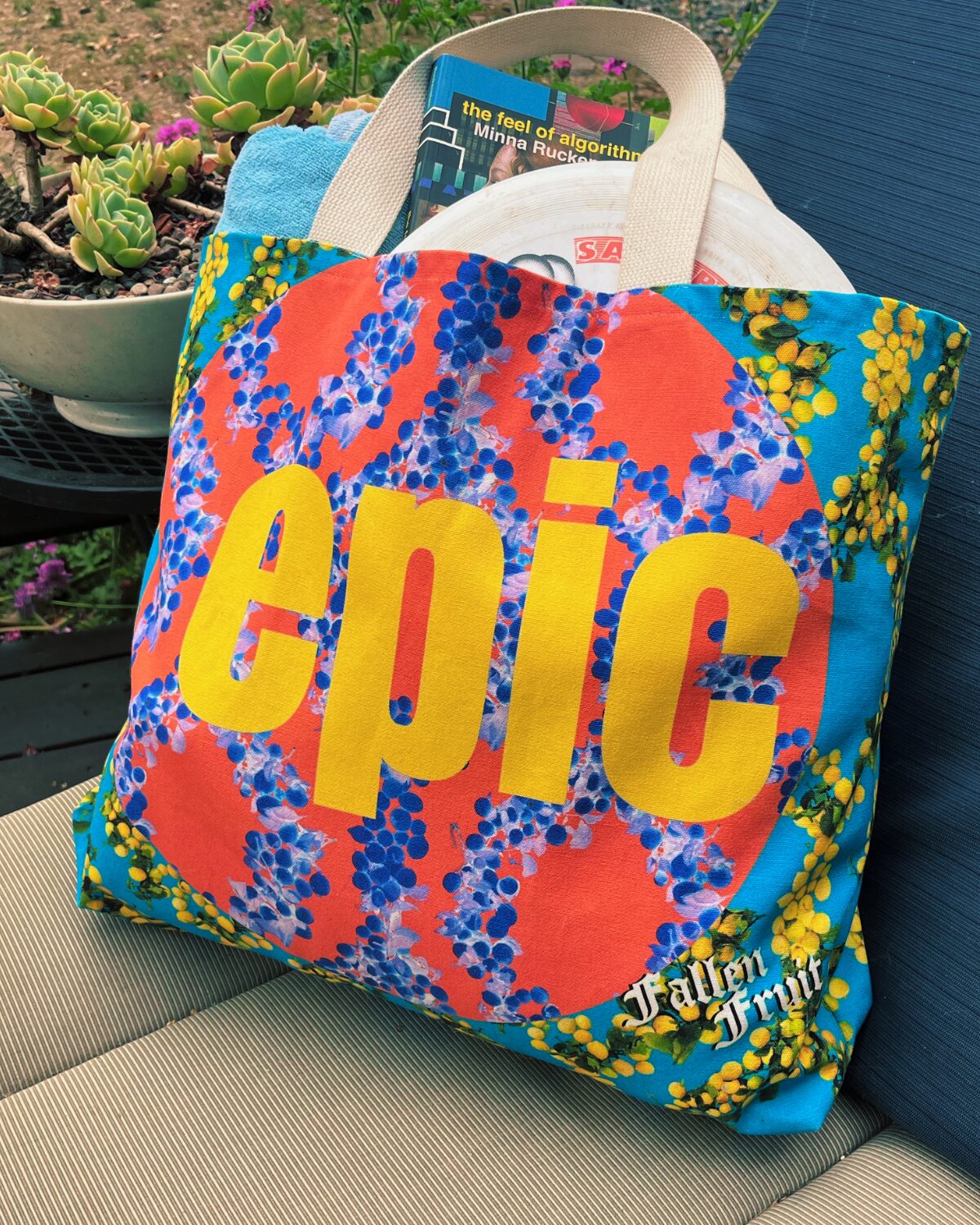 Photo of the Lemon Tote Bag. The bag is a vibrant blue with a pattern of strings of lemons, and a large EPIC logo is a circle indicated by changing of the pattern colors and large yellow letters 'epic' in the middle. In the bag are beach towels, a frisbee, and a copy of the book The Feel of Algorithms.