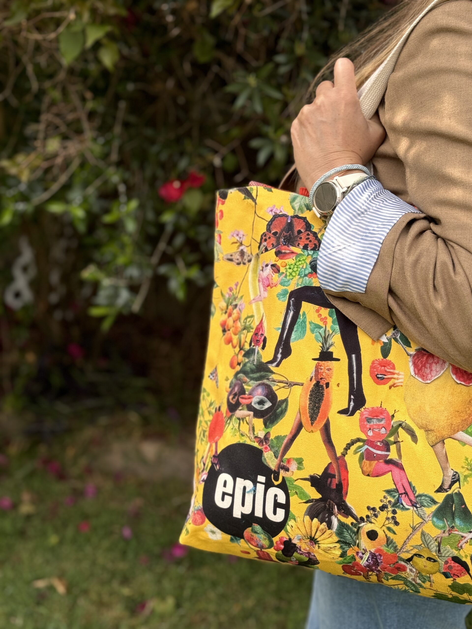 Close-up photo of someone with a tote bag over their shoulder. The bad is yellow with cut-out photos of people and fruit combined in collage style. In many cases a piece of fruit appears to be a body, with human legs. The EPIC logo is in the bottom left corner; a black circle with 'epic' in which letters in the middle.