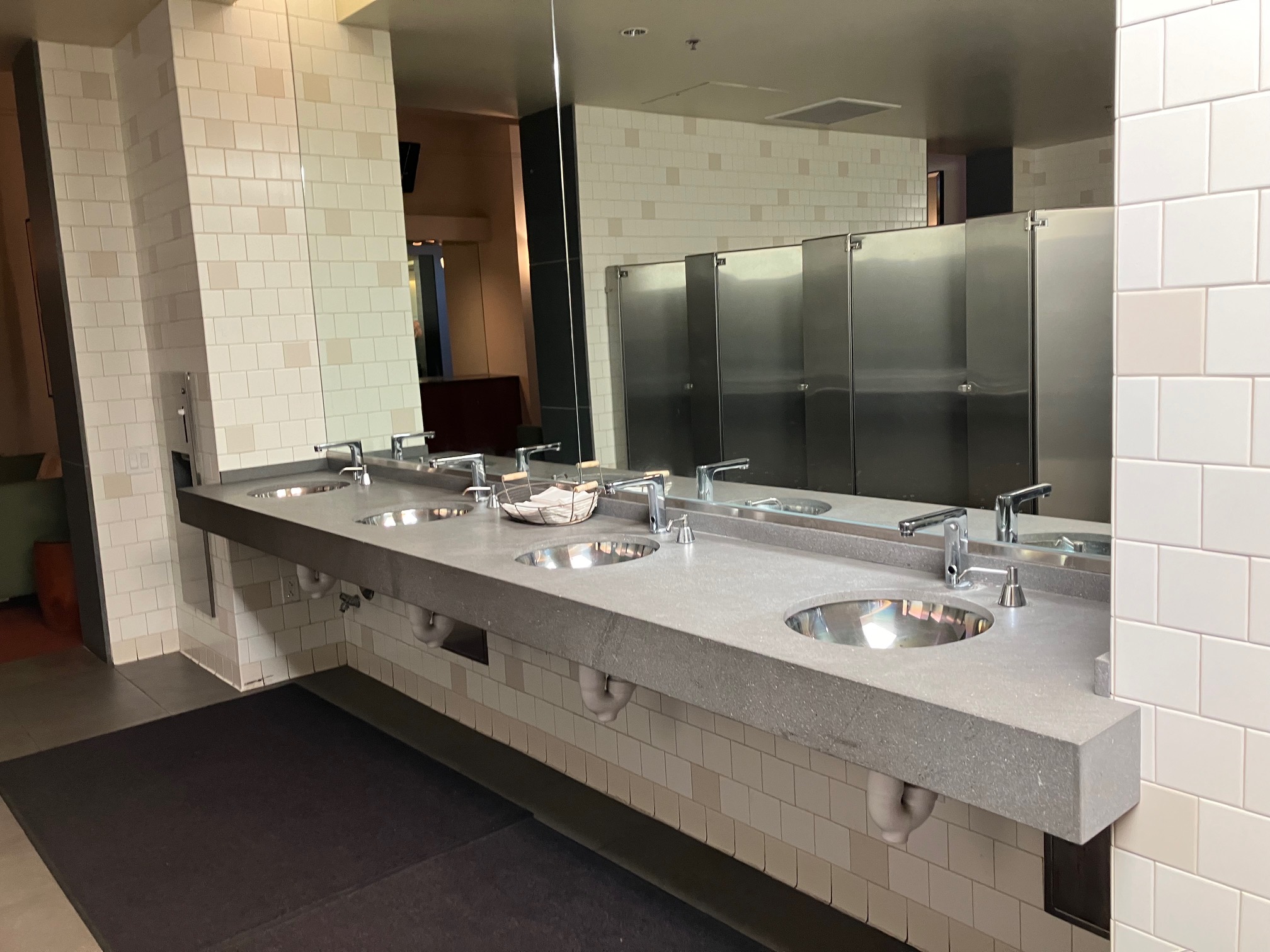 One of the bathrooms is marked with a sign saying 'women' though all conference-goers are welcome to use it.  There are multiple stalls with an accessible stall at the far end of the bathroom. There is a countertop with 4 sinks in a row.  The sinks have automatic taps and each has a pump soap dispenser to the right. There are paper towels in a basket in the middle of the sink area, and a wall-mounted trash bin is on the left side of the sinks. Inside the accessible stall there are two wall-mounted grab rails: one on the lefthand wall and one behind the toilet. 