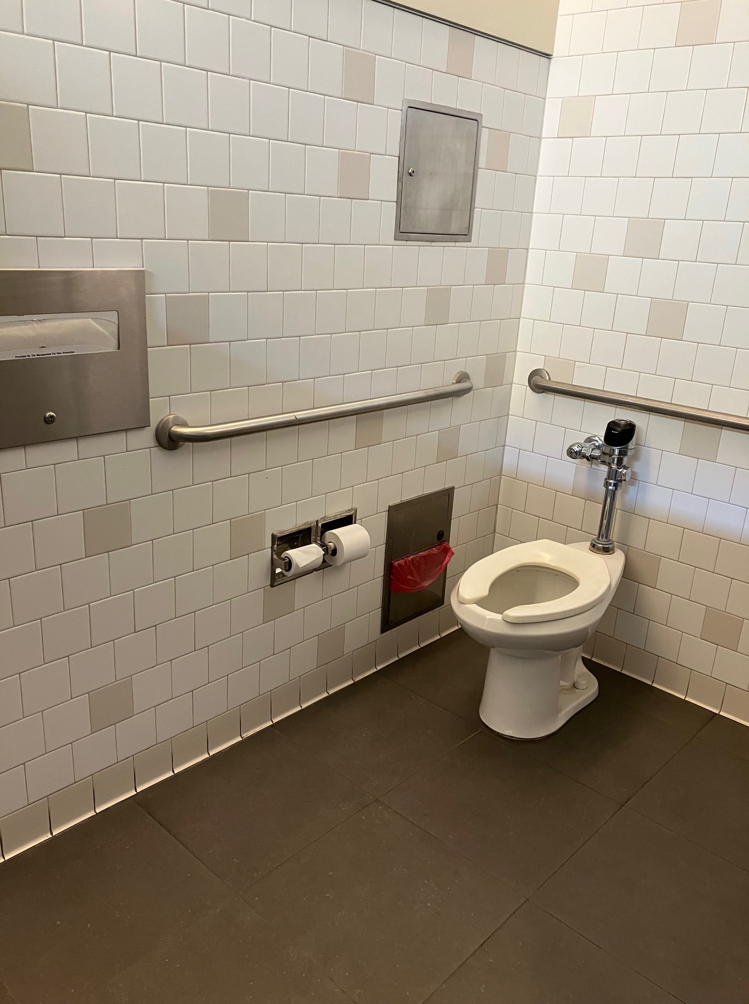 One of the bathrooms is marked with a sign saying 'women' though all conference-goers are welcome to use it. There are multiple stalls with an accessible stall at the far end of the bathroom. There is a countertop with 4 sinks in a row. The sinks have automatic taps and each has a pump soap dispenser to the right. There are paper towels in a basket in the middle of the sink area, and a wall-mounted trash bin is on the left side of the sinks. Inside the accessible stall there are two wall-mounted grab rails: one on the lefthand wall and one behind the toilet.